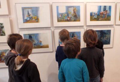 Kinder besuchen die Ausstellung “Über Tisch und Bänke” bei LesArt. Sie betrachten Bilder an den Wänden.