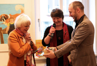 Der LesArt-Mitarbeiter Frank Kurt Schulz übergibt der Künstlerin Ilon Wikland ein Geschenk anlässlich der Ausstellungseröffnung von “Über Tisch und Bänke”.