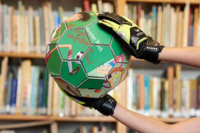 Ein mit dem farbigen Buchcover von Karsten Teichs “Wie Franz den Fußball erfand” bedruckter Fußball aus Pappe wird von Kinderhänden in Torwandhandschuhen gehalten