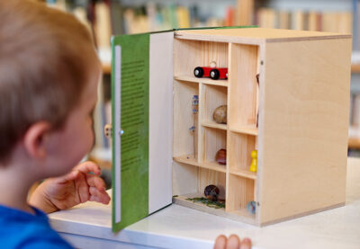 Ein Kind öffnet die Tür eines hölzernen Buchobjektes. Dahinter: Setzkasten mit rotem Spielzeugauto, Schneckenhäusern, Lavendelzweig und Spielfigur.