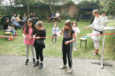 Anlässlich der Ausstellungseröffnung AUSERLESENES zerschneiden Kinder feierlich rotes Band im Eingangsbereich des LesArt-Gartens.