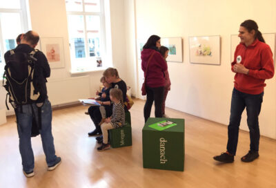 Erwachsene und Kinder besuchen die Ausstellung AUSERLESENES. 30 Jahre LesArt bei LesArt. An den Wänden hängen Erzählbilder. Im Raum stehen grüne Hocker in Würfelform, deren Seiten mit verschiedenen Wörtern bedruckt sind