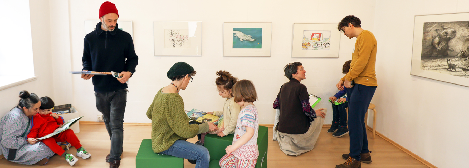 Erwachsene und Kinder besuchen die Ausstellung “AUSERLESENES. 30 Jahre LesArt” und betrachten Bücher. Sie sitzen am Boden, oder auf grünen Hockern in Würfelform.