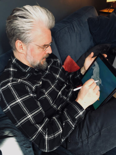 Illustrator Martin Baltscheit sitzt auf dunklem Sofa und zeichnet sein Erzählbild mit Krokodil auf einem Grafiktablet. Er trägt Brille, hat graues Haar, einen Vollbart und ist leger in schwarzer Hose und dunklem Karohemd gekleidet.